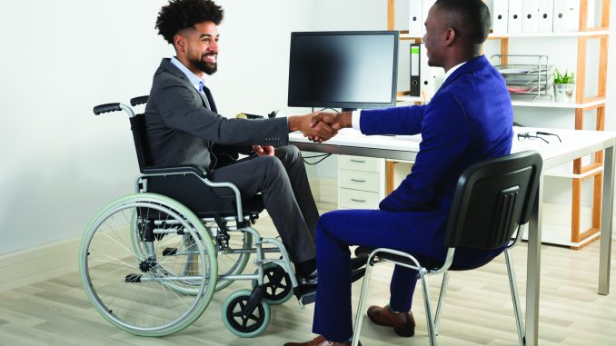Two professionals meeting in an office, one using a wheelchair, shaking hands across a desk during what appears to be an interview or business meeting.
